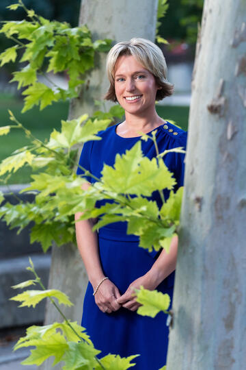 Alison Sloat wearing a blue dress partially behind a tree trunk with large green leaves in front of her