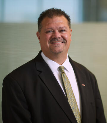 Brian Labus smiling and wearing a black suit jacket, white collared shirt, and light colored tie
