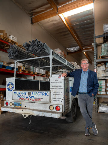 Wearing a blue checkered blazer, blue checkered dress shirt and gray pants, Joe Murphy, winner of the 2024 Silver State Award, leans his right arm on a Murphy Electric work truck parked in the shop of Murphy Electric