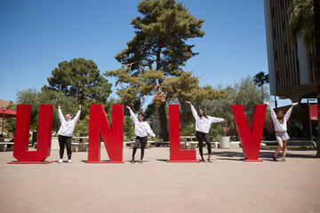 Four students create a U, N, L, and V with their arms interspersed between tall red UNLV letters